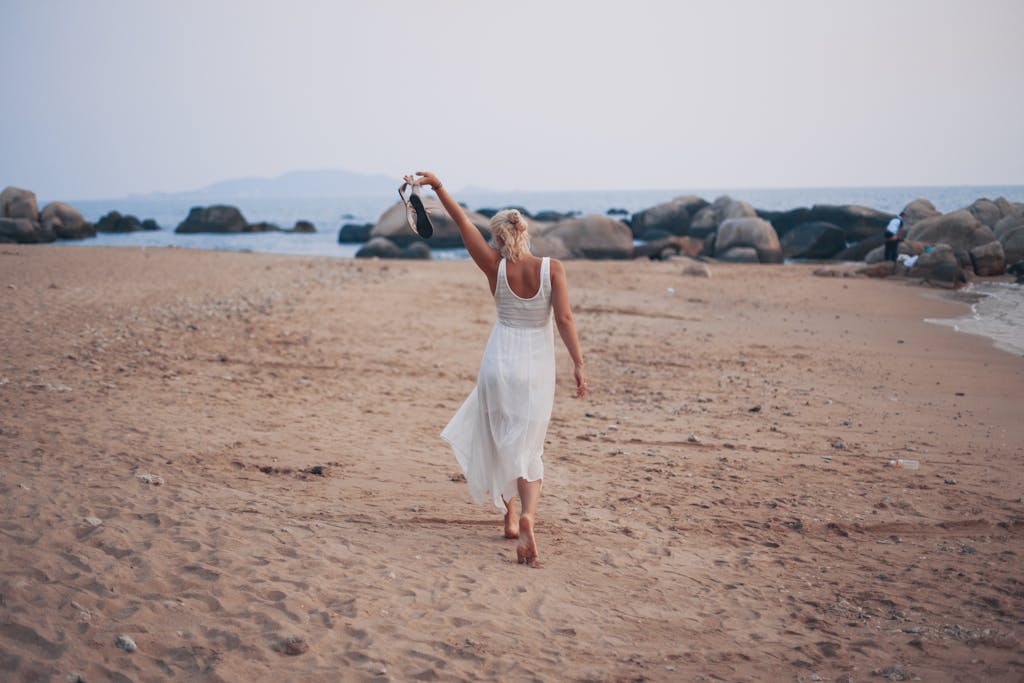 A woman in a white dress walks barefoot on a scenic beach, symbolizing freedom and relaxation.