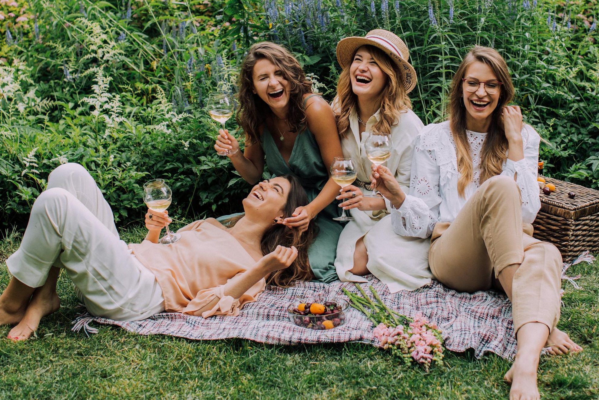 Four women enjoying a picnic, laughter, and wine in a vibrant summer garden.